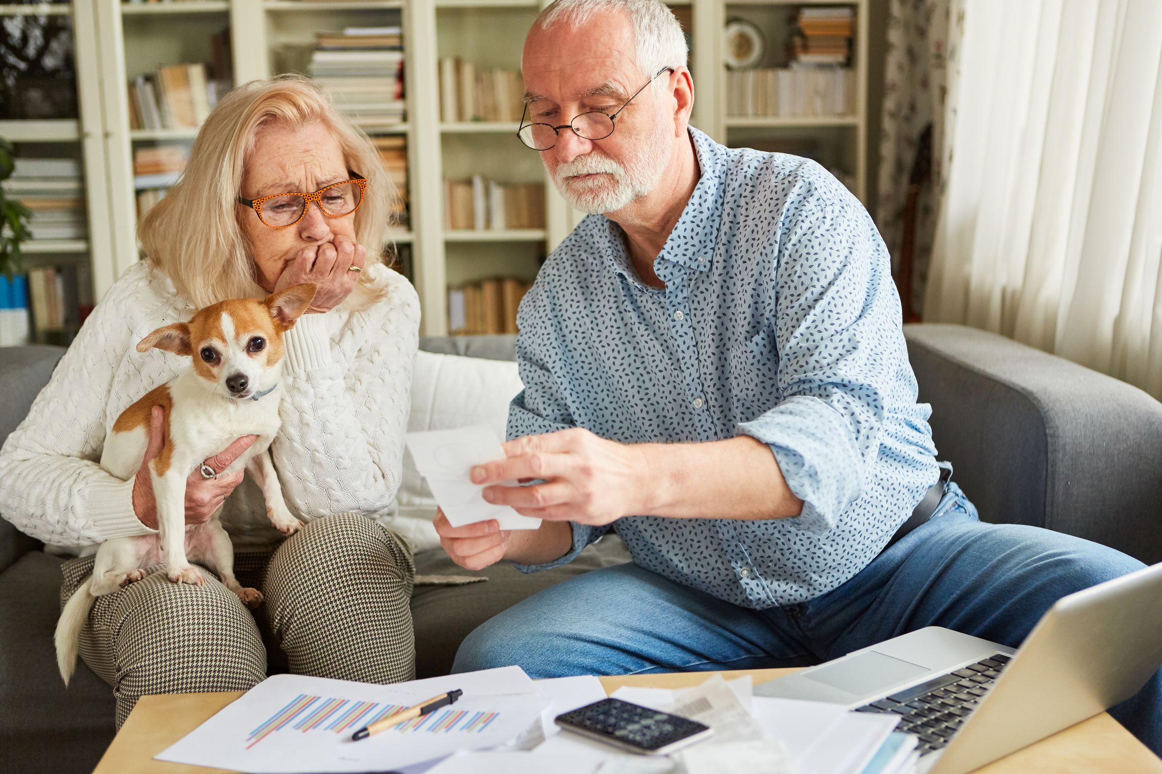 Senior couple sit on sofa at home with their dog, looking worried as they sort through bills.